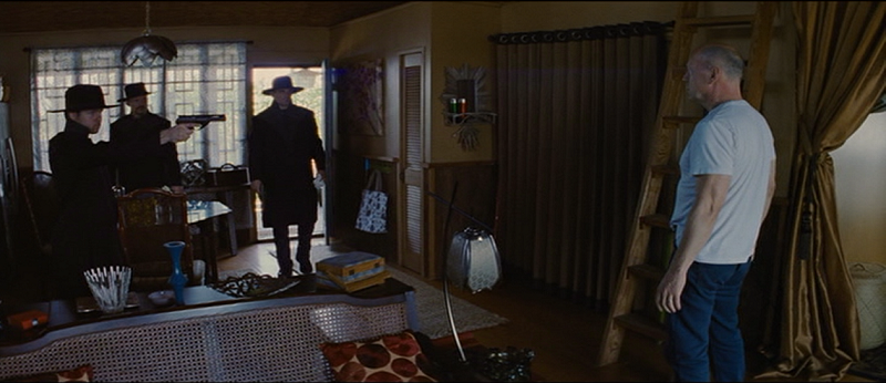 Three men in black coats and wide-brimmed hats enter Joe's home. One points a long-barreled gun at Joe, who stands off to the right.